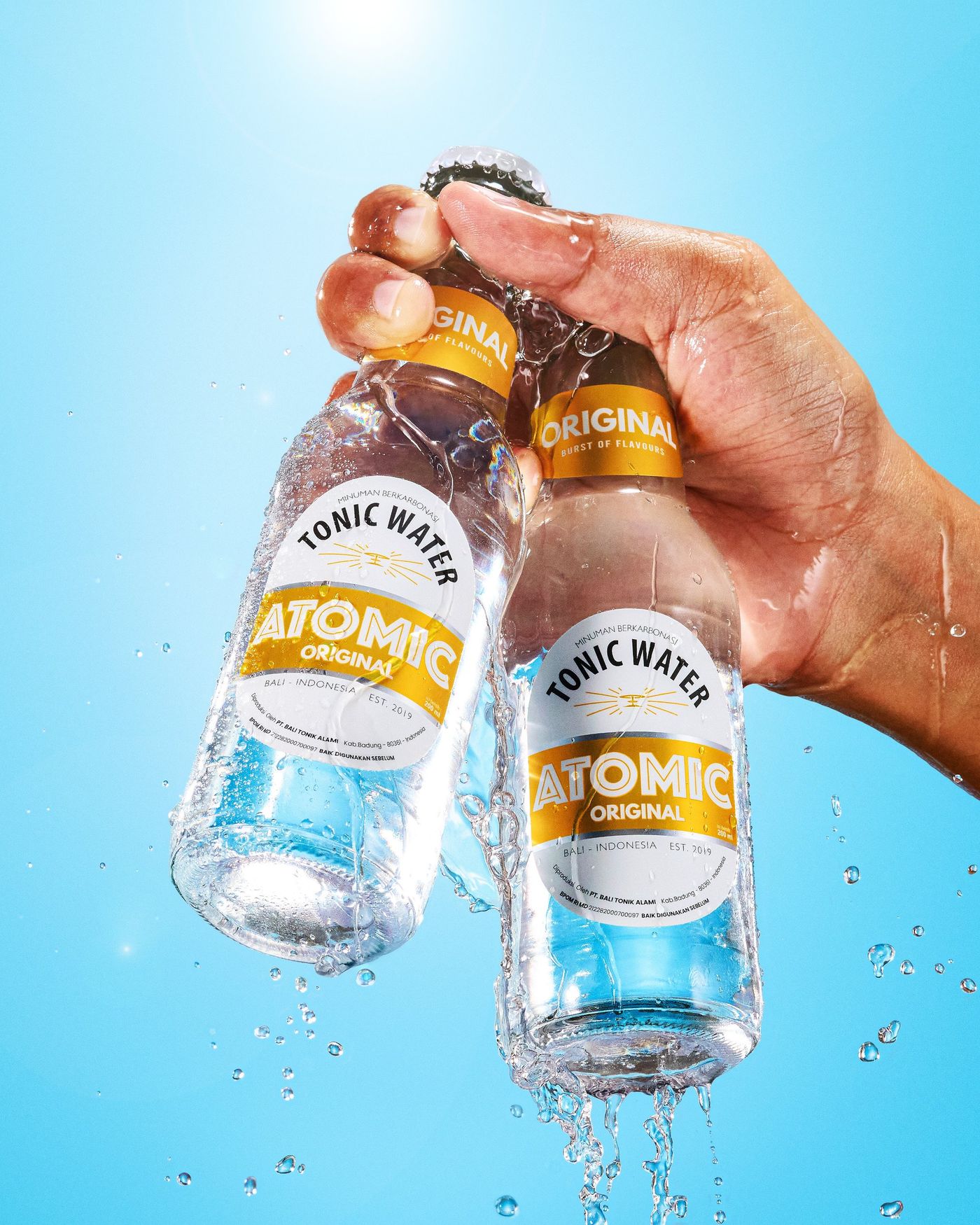 A hand holding two bottles of Atomic Original Tonic Water with a splash, against a blue sky